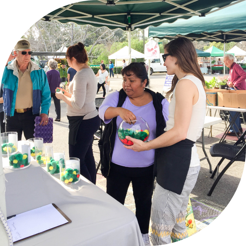 Cristelle is at a farmer's market inviting community members to vote on budget items that are important to them.