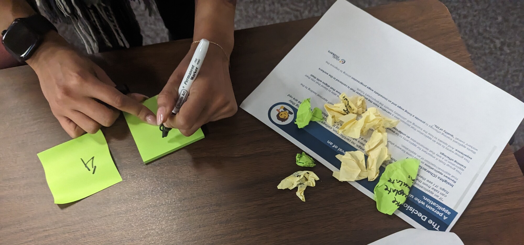 A session attendee's hands can be seen writing on a post-it. They have an open packet of background info in front of them, as well as a few crumpled up post-its.