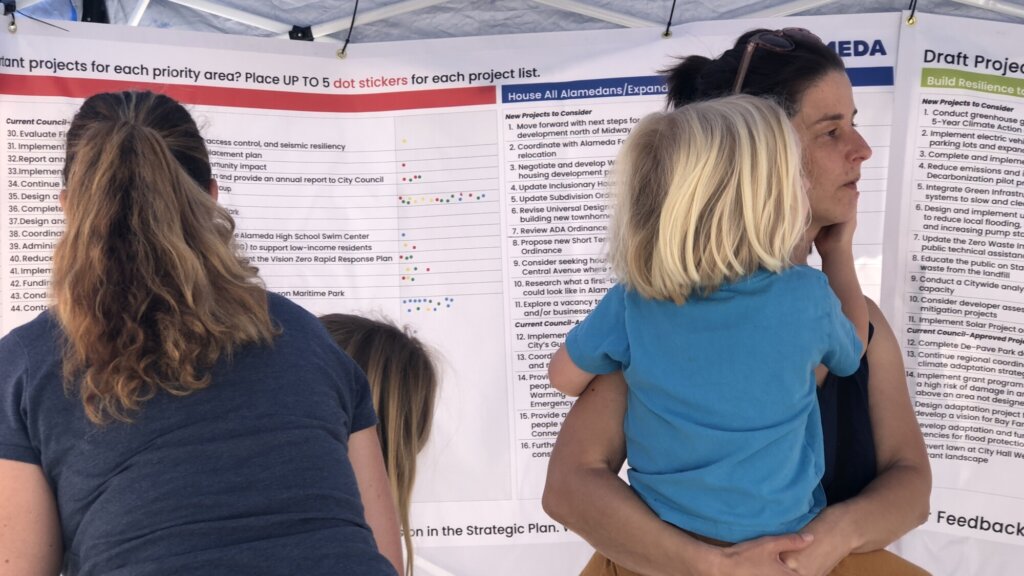Two adults and two children are looking at and in front of two three large pieces of paper. They are in a large foldable tent or shade structure at the Alameda farmer's market. The pieces of paper are hung up on three sides of the tent. The paper has potential details and things to vote on that they'd like to see in the plan. They are allowed to place dots next to the statements they like.