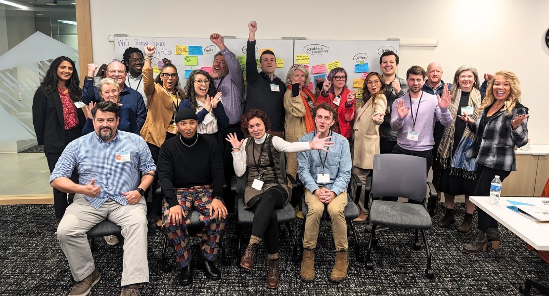 Fellows and Friends of the Workforce Transformation Corps are grouped together, smiling and waving, in front of a whiteboard of post-its.