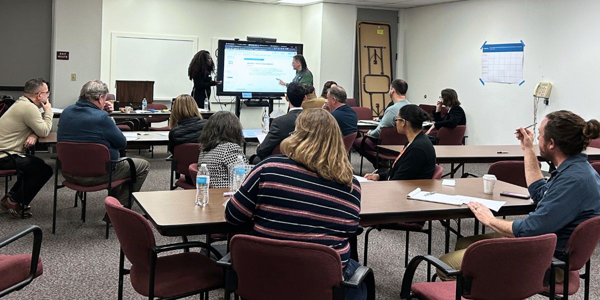 In person ideation session. Brit and Eva stand at the front of the room, pointing at a slide on the tv. Participants are seated around different tables listening to instructions on the ideation exercise.