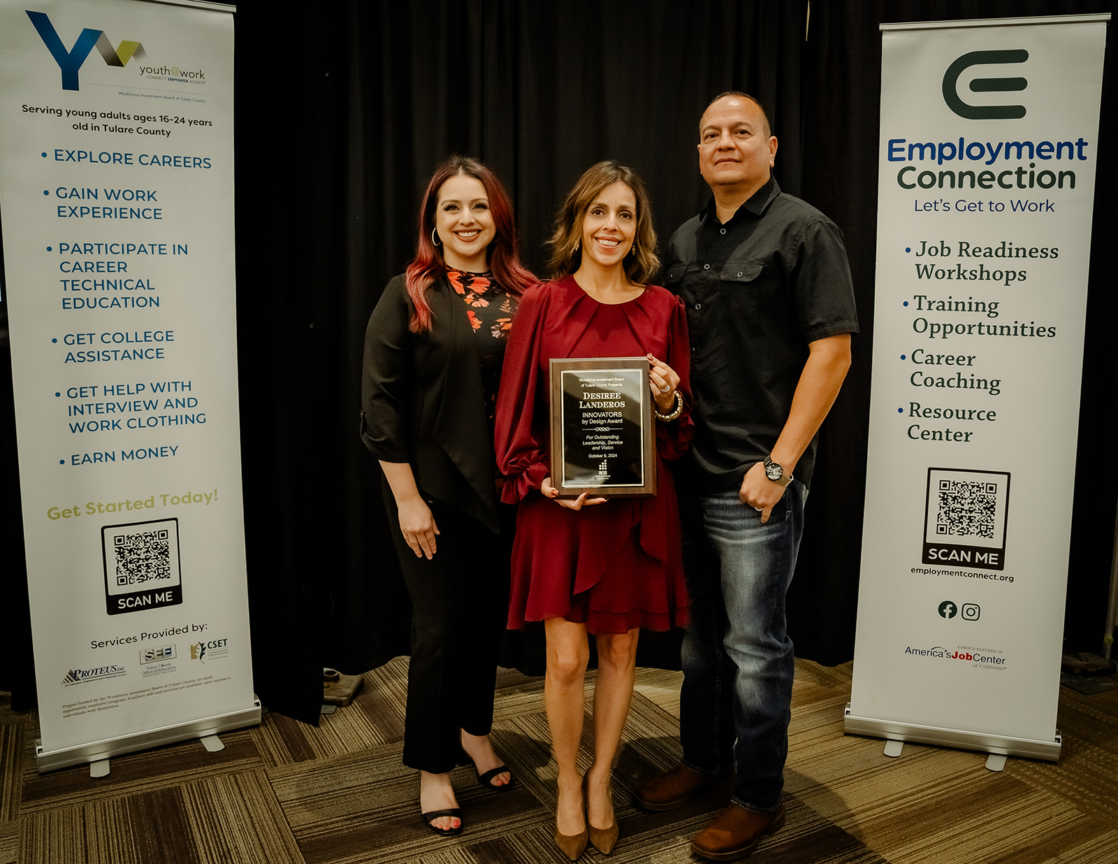 Desiree Landeros stands in front of conference banners, holding the plaque that she was awarded as one of 2024's Workforce Professionals of the Year. Beside her are her adult daughter and husband.