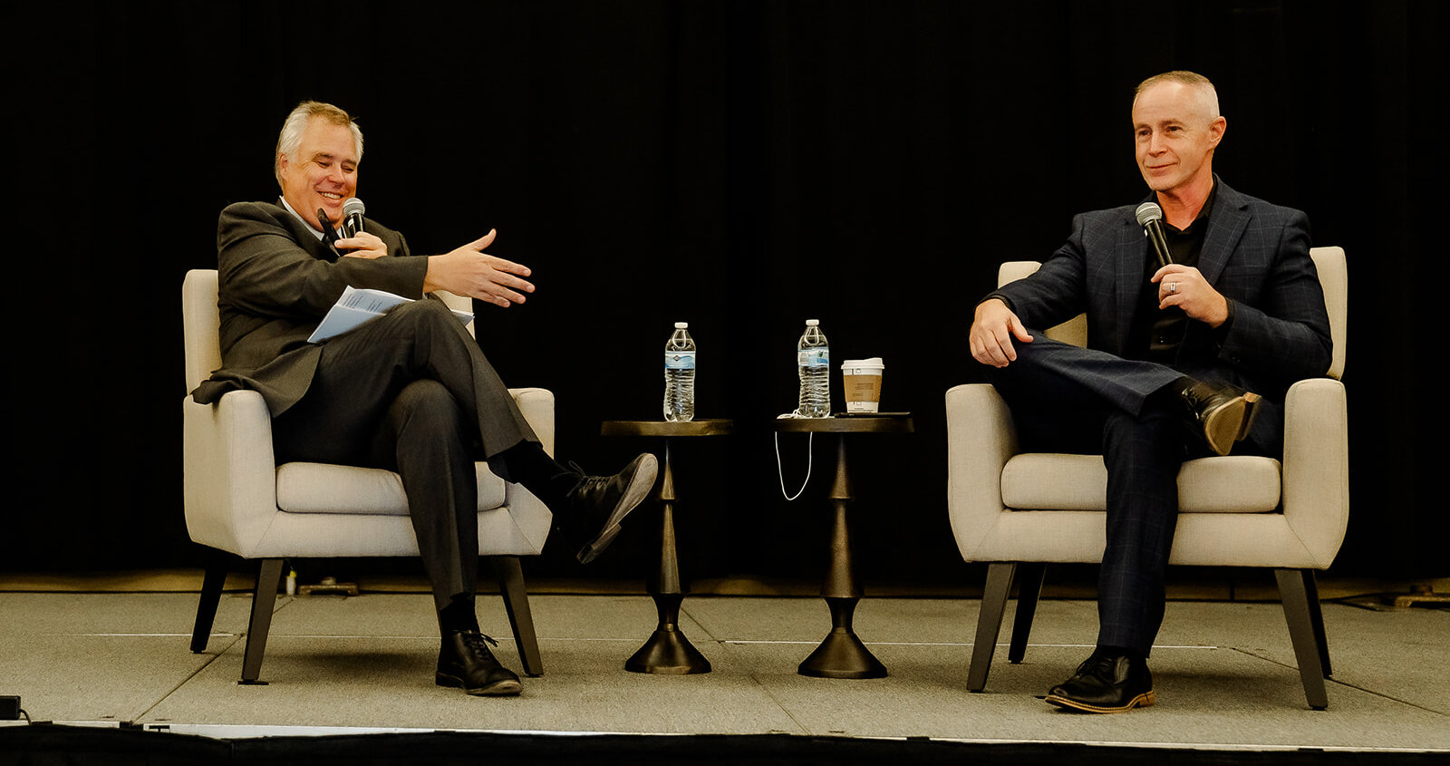 Adam Peck & Stewart Knox are seated in chairs on stage, in a fireside-style conversation.