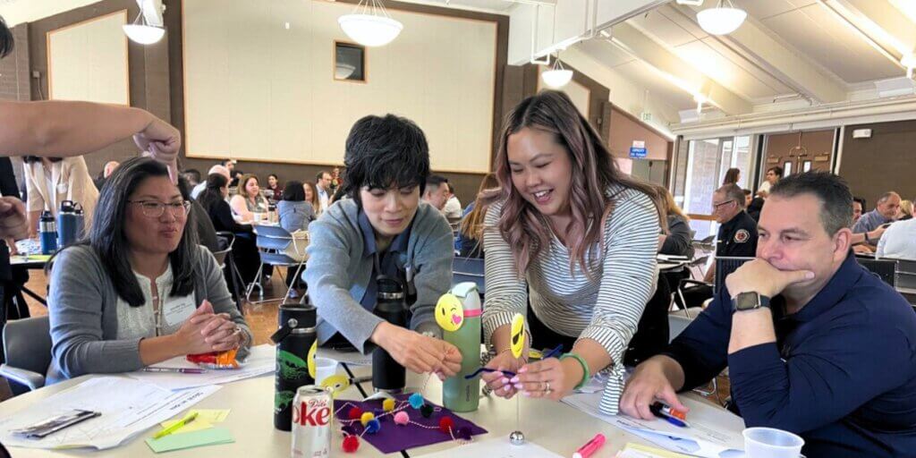 In this picture, four people are grouped around a table. Two women in the middle are half standing up, arranging a prototype made of crafting supplies, while others around the table look on or offer directions.
