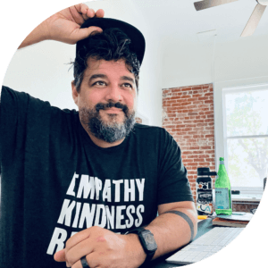 Headshot of Tom Price. He is looking off camera, with a hand on his cap and an arm resting on his desk. He's wearing a black t-shirt that shows the words 'empathy' and 'kindness'.