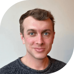 Headshot of James wearing a gray sweater and standing in front of a beige background. He has short, dirty blonde hair and blue eyes.