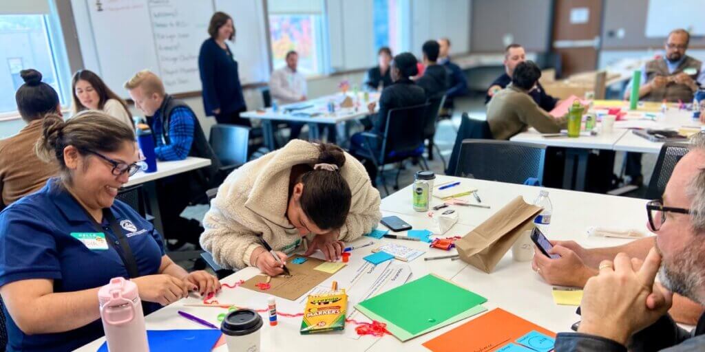 Picture from a Learning Lab Session. There's a conference room with several tables of different teams. One team, near the camera can be seen building prototypes with craft supplies. One person is heads down, deep in focus on what they are drawing/writing.