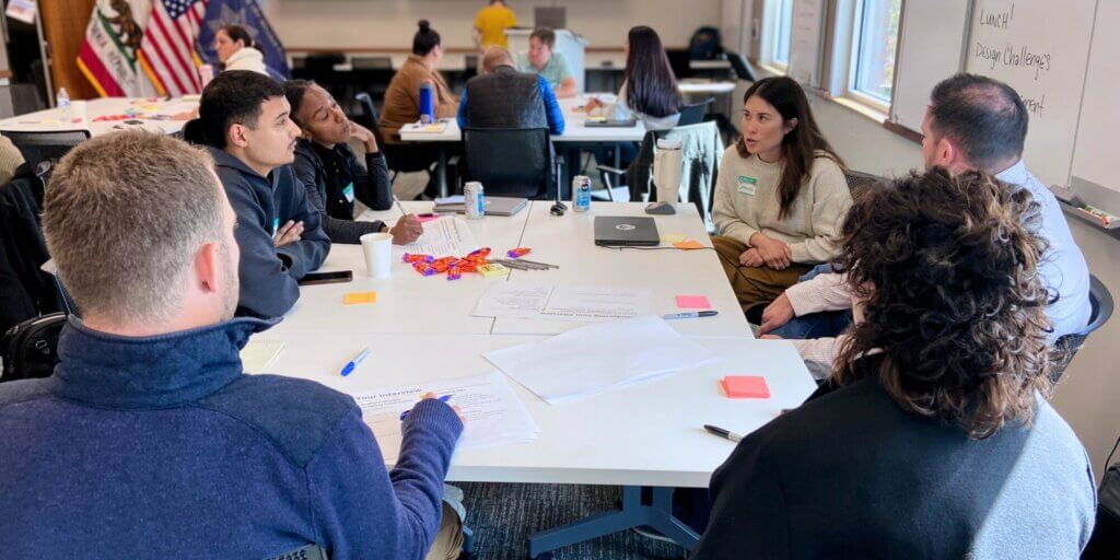 A small team is seated around a table and taking notes as one person is being interviewed.