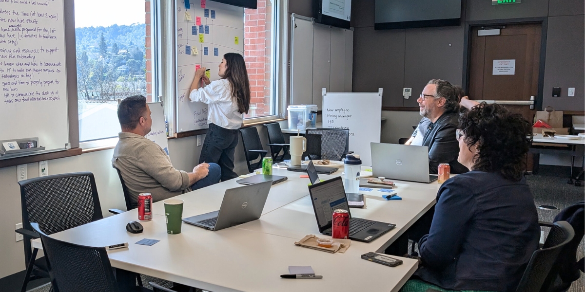 A team of four are working around a table. One person is standing and writing on a post-it, which is surrounded by other post-its that have already been added. Her three team members are all watching from where they are seated, with laptops available nearby.