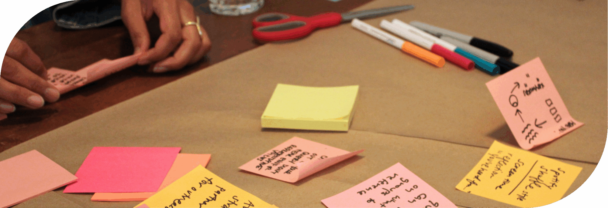 A close-up of several post-its with ideas and drawings arranged on a roll of brown paper on a table. They are seen from upside down in the photo as the photographer stands on the other side of the table. In the top left corner, a pair of hands are holding a few post-its, about to add them to the group. There are also several colored markers and sharpies nearby.