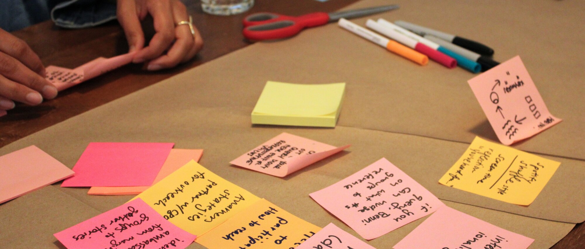 A close-up of several post-its with ideas and drawings arranged on a roll of brown paper on a table. They are seen from upside down in the photo as the photographer stands on the other side of the table. In the top left corner, a pair of hands are holding a few post-its, about to add them to the group. There are also several colored markers and sharpies nearby.
