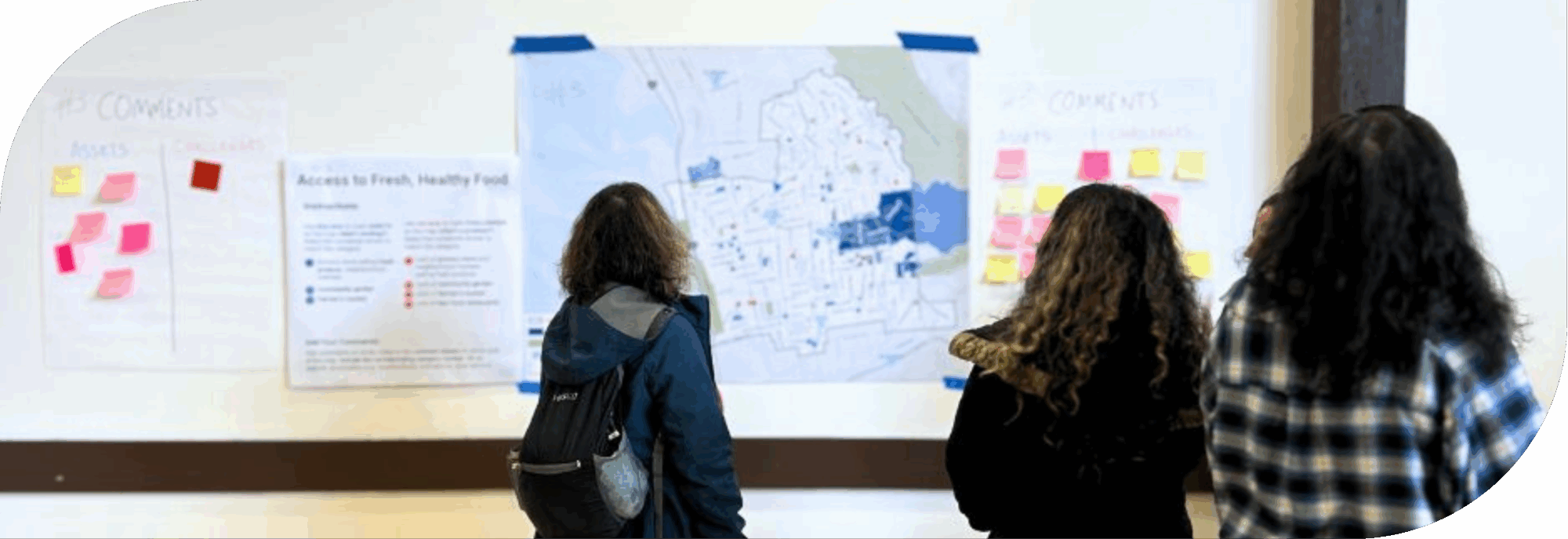 Three people are standing in front of a wall with several posters taped up. The posters include a large map with red and blue dots placed in different locations, there are also a couple posters where people have already placed several post-its.
