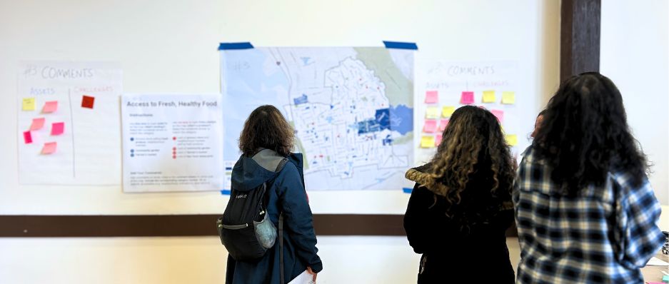 Three people are standing in front of a wall with several posters taped up. The posters include a large map with red and blue dots placed in different locations, there are also a couple posters where people have already placed several post-its.