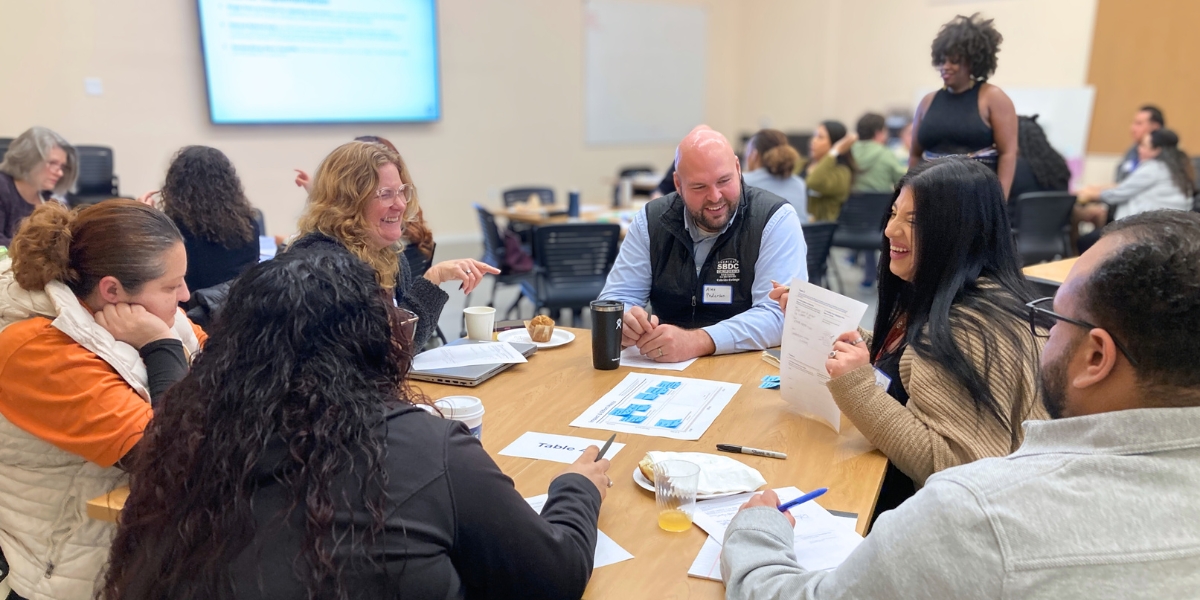 A small group, seated around the table, is laughing together as one person is showing something from their worksheet.