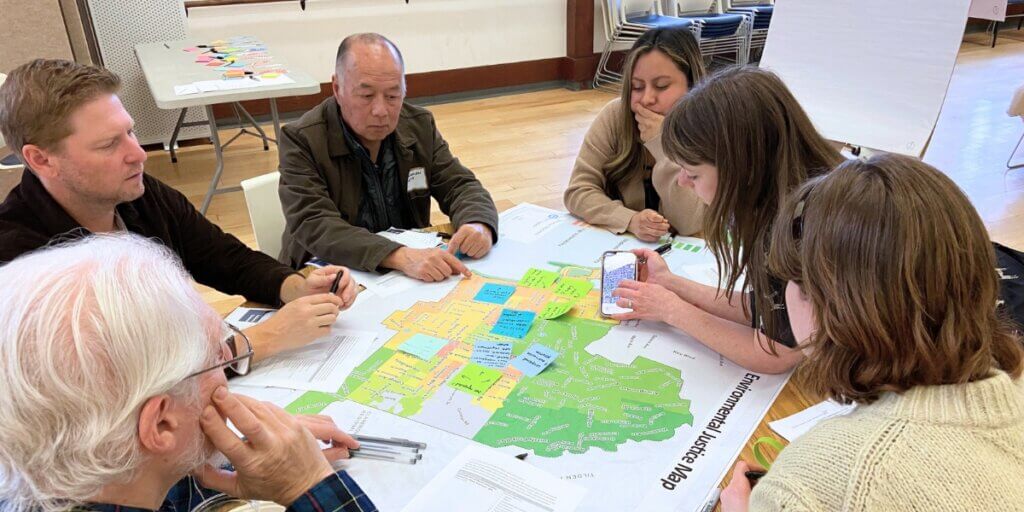 Six people are seated around a table. In the middle is a large map of the City of Berkeley with some areas in green, yellow, or orange. On top of the map are several post-its. A facilitator is leaning in showing people something on their phone as part of the discussion.